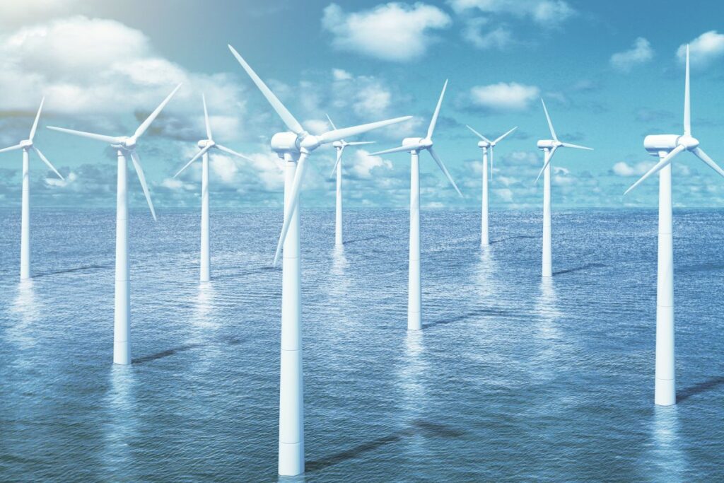 Offshore wind turbines standing in the ocean under a bright blue sky with scattered clouds. The windmills rise from the water’s surface, representing large scale offshore wind energy and renewable power generation.