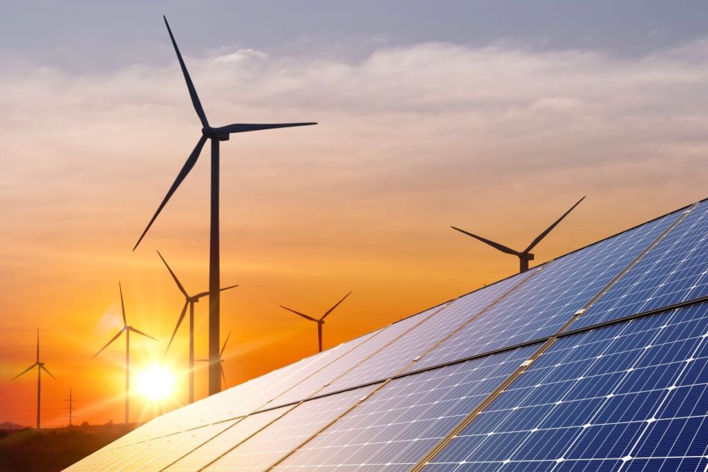 Wind turbines rising behind rows of solar panels at sunset, with the sun glowing low on the horizon. The combination of wind and solar infrastructure highlights a clean energy concept and the transition to renewable power generation.