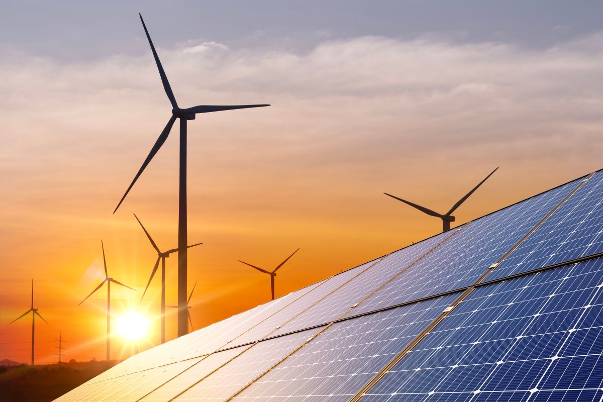 Wind turbines rising behind rows of solar panels at sunset, with the sun glowing low on the horizon. The combination of wind and solar infrastructure highlights a clean energy concept and the transition to renewable power generation.