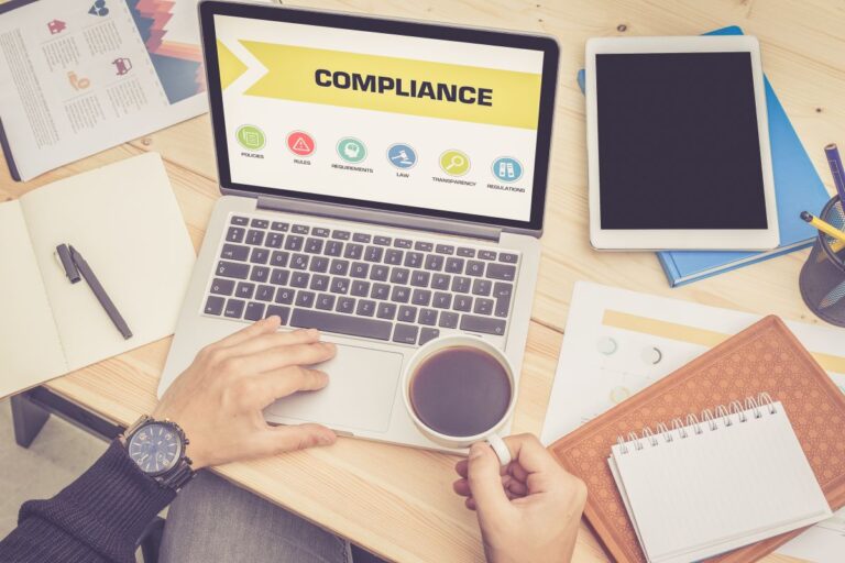 Laptop screen displaying the word COMPLIANCE with icons labeled policies, rules, requirements, law, transparency, and regulations. A person holds a cup of coffee while working at a desk with a tablet, notebook, and documents, emphasizing regulatory compliance and business governance.