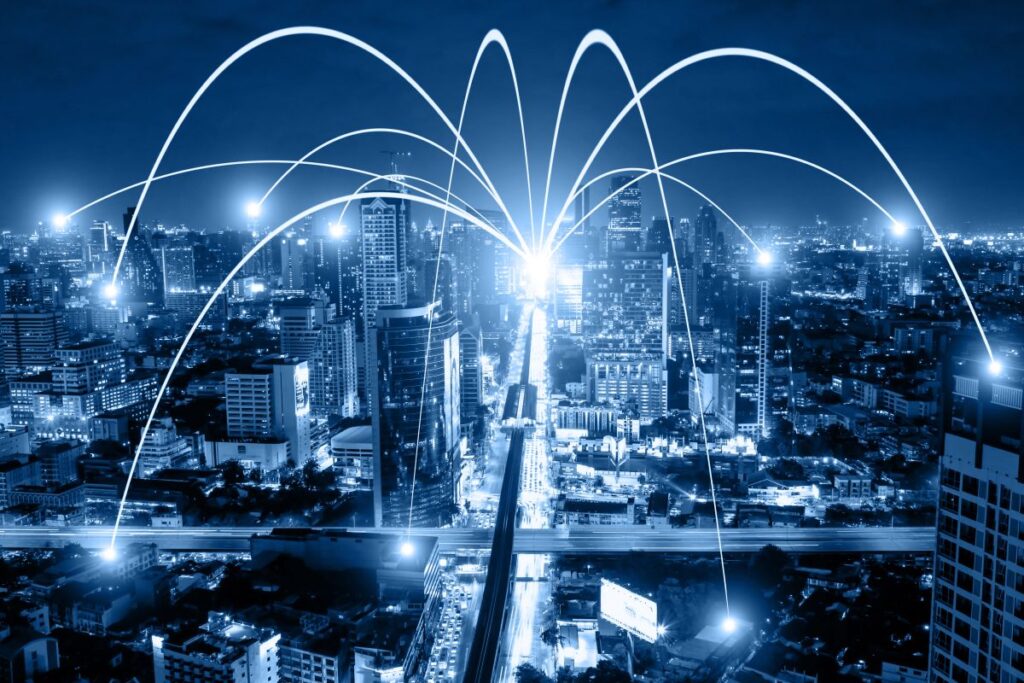 Night city skyline with glowing connection lines arcing between buildings to represent business networking and WiFi communication technology. The illuminated urban landscape and digital links highlight smart city infrastructure and grid digitization.