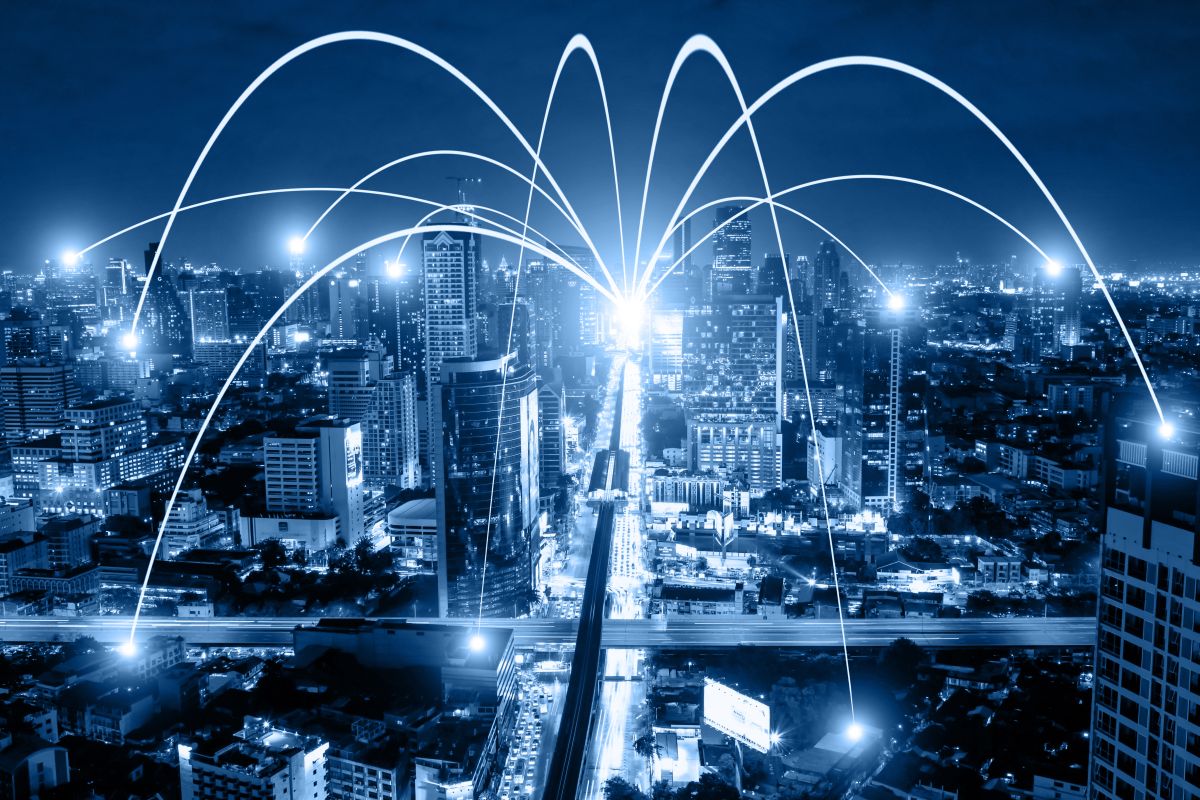 Night city skyline with glowing connection lines arcing between buildings to represent business networking and WiFi communication technology. The illuminated urban landscape and digital links highlight smart city infrastructure and grid digitization.