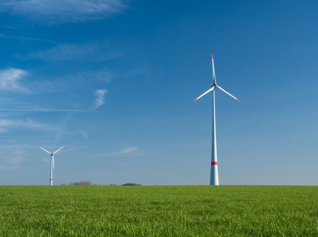 Two wind turbines standing on a green meadow under a clear sunny blue sky. The tall white wind power plants highlight clean renewable energy generation in a rural landscape.
