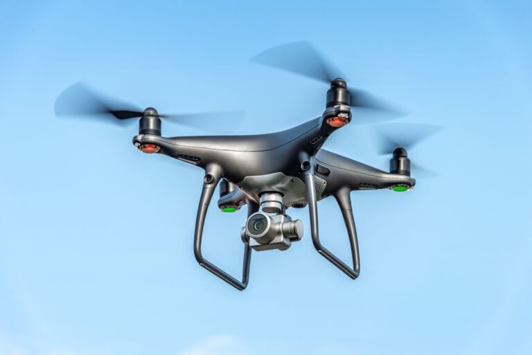 Quadcopter drone flying in a clear blue sky with four spinning propellers and a mounted camera underneath. The unmanned aerial vehicle is captured midair, representing modern drone technology used for security and aerial surveillance.
