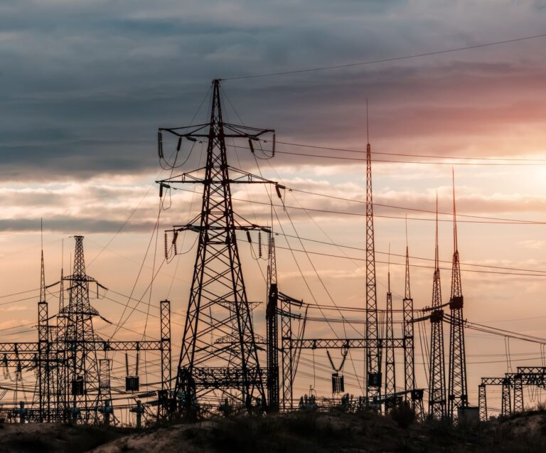 Distribution electric substation with power lines, lattice towers, and transformers silhouetted against a sunset sky. The network of transmission structures highlights critical grid infrastructure supporting electricity distribution and substation security.