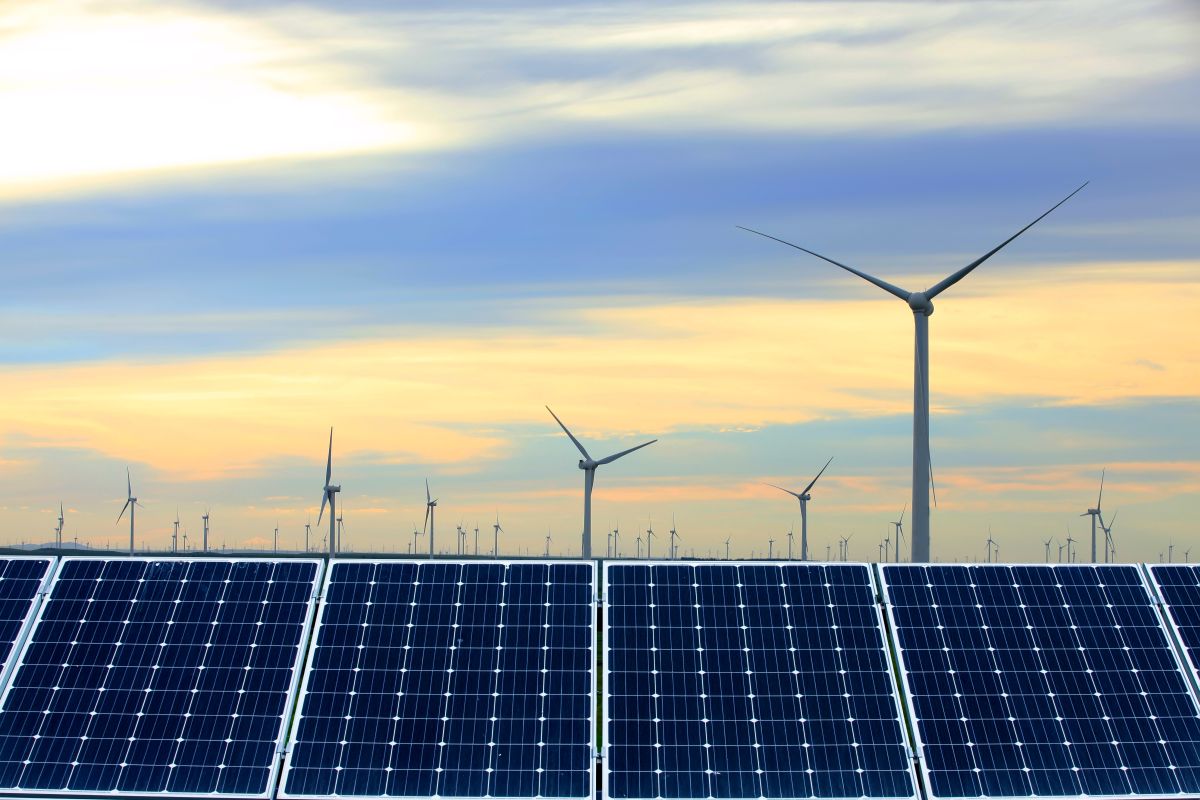Solar photovoltaic panels in the foreground with multiple wind turbines generating power under a colorful sunset sky. The image represents a renewable energy concept combining solar and wind resources for distributed energy generation.