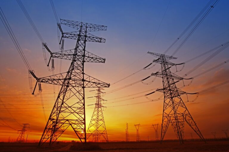 WireElectricalEnergyAtSunset Silhouettes of high voltage transmission towers and power lines stretching across the horizon at sunset. The orange and blue sky backlights the electrical grid infrastructure, emphasizing energy distribution during peak demand hours.