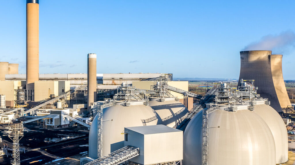 Industrial carbon capture and storage facility with large domed tanks, smokestacks, and cooling towers at a power plant site. The complex infrastructure represents carbon capture and storage technologies used to reduce industrial emissions.