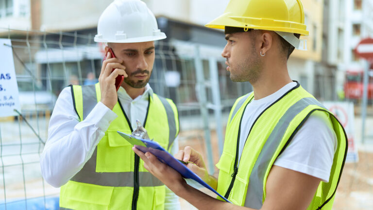 Two construction workers wearing hard hats and high visibility vests review paperwork on a clipboard at an active job site while one speaks on a phone. The image reflects preparation and documentation processes involved in a NERC audit.