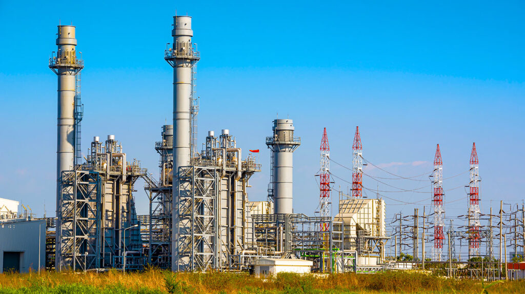 Industrial power generation facility with tall exhaust stacks, complex piping systems, and adjacent high voltage transmission towers under a clear blue sky.