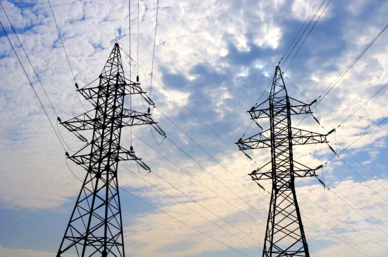 Two electricity pylons with multiple power lines stretching across a partly cloudy sky. The tall transmission towers stand against soft blue and white clouds, emphasizing large scale power grid infrastructure.