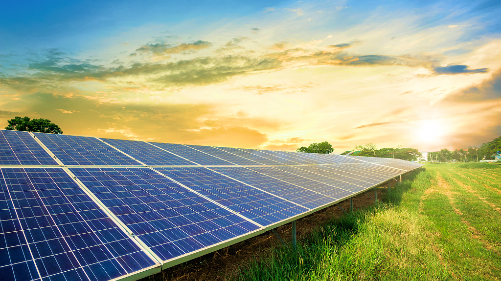 Wide view of a solar panel array installed on a grassy field at sunset, with golden light illuminating the photovoltaic modules. The clean energy landscape represents grid connected renewable resources and compliant integration of inverter based resources within modern power systems.