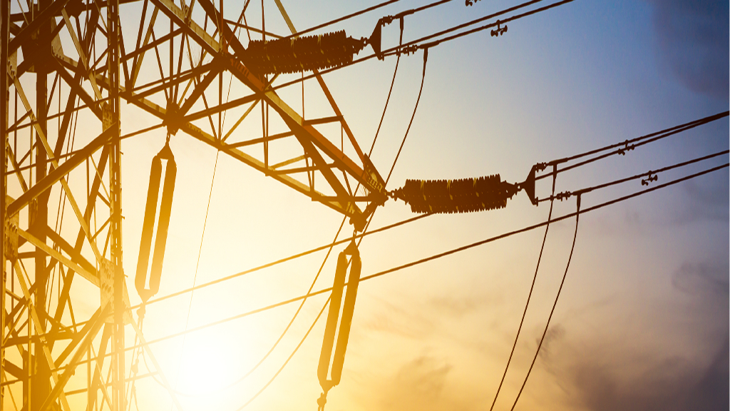 Close-up of a high-voltage transmission tower with detailed view of insulators and power lines, set against a dramatic sunset sky. The golden light and structural focus emphasize energy infrastructure, aligning with FAC-008 compliance and transmission reliability topics.