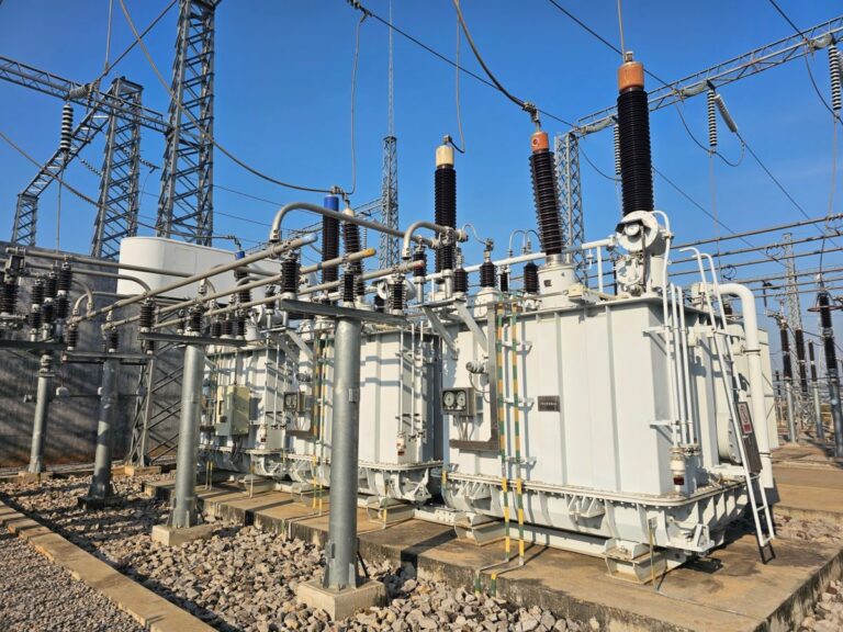 Three single phase power transformers installed in an outdoor switchyard with high voltage bushings and overhead transmission lines. The large electrical equipment and steel lattice structures highlight critical substation infrastructure supporting the power grid.