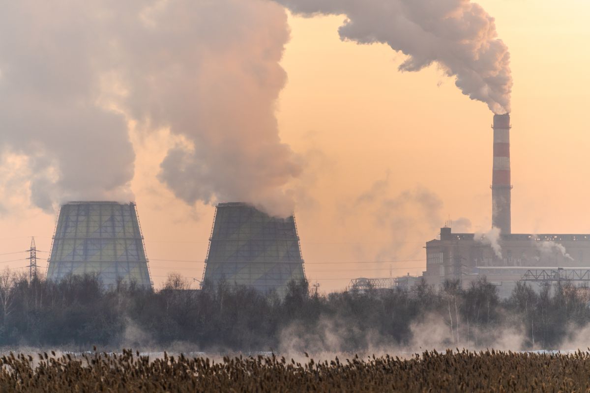 Industrial power plant in winter with large cooling towers and a tall smokestack releasing heavy smoke into the cold air. Steam and emissions rise above a frosty landscape, emphasizing industrial activity and air pollution during winter conditions.