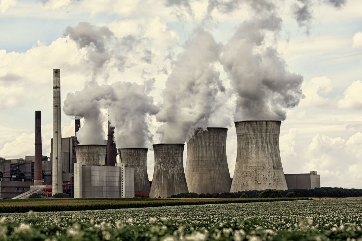 Coal power plant with multiple large cooling towers and smokestacks releasing thick plumes of smoke into the sky. The industrial facility stands beyond an open field, highlighting fossil fuel energy production and air emissions.
