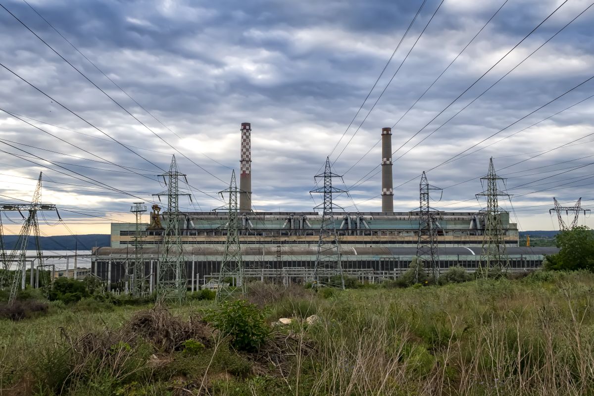 A thermal power station with smoke rising from chimneys, surrounded by power lines, buildings, and green trees under a cloudy sky.