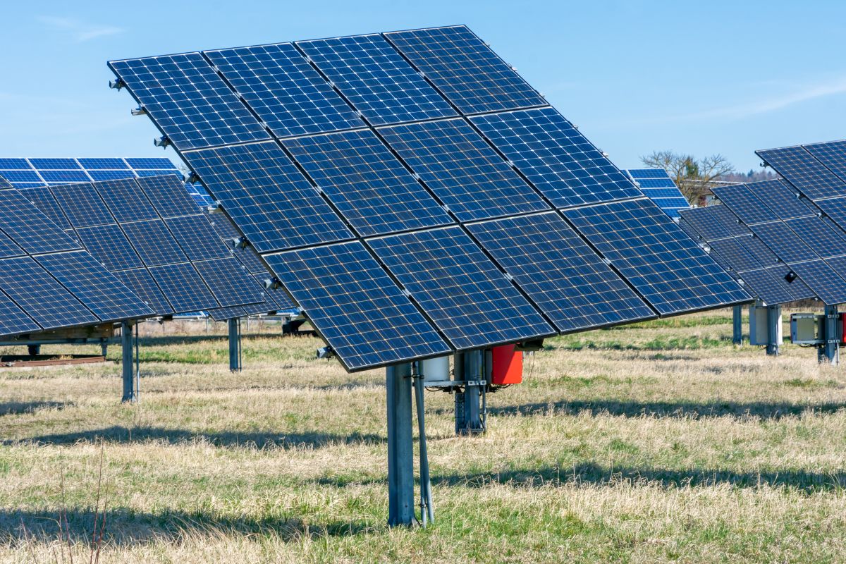 A row of solar panels in a field, angled towards the sun, with electrical equipment visible at the base of each panel.