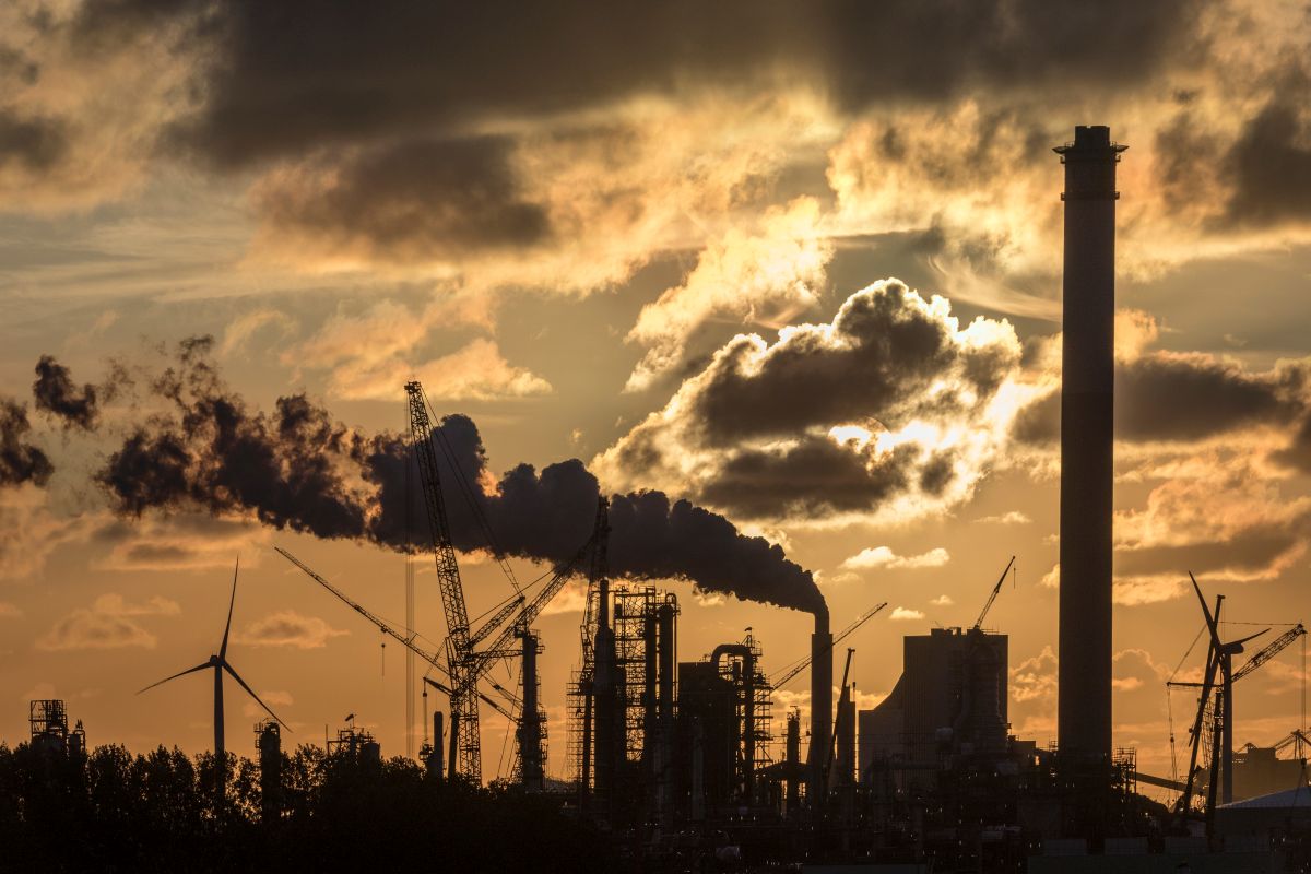 Industrial Sunset - Rotterdam - Holland A dramatic sunset over an industrial landscape in Rotterdam, Holland, showcasing smokestacks emitting clouds of smoke, towering cranes, wind turbines, and other industrial infrastructure. The sky is filled with dark clouds, creating a striking contrast between the warm sunset and the industrial scene.