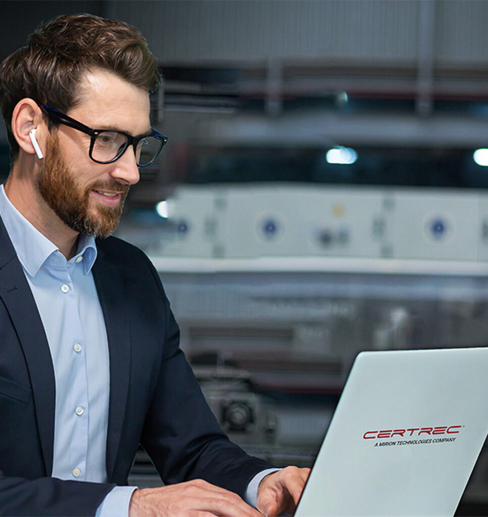 Professional man with a beard and glasses wearing a navy blazer and white shirt, using wireless earbuds while looking at something offscreen. The background shows a blurred industrial or control room environment, relating to NRC fatigue rule compliance in the workplace.