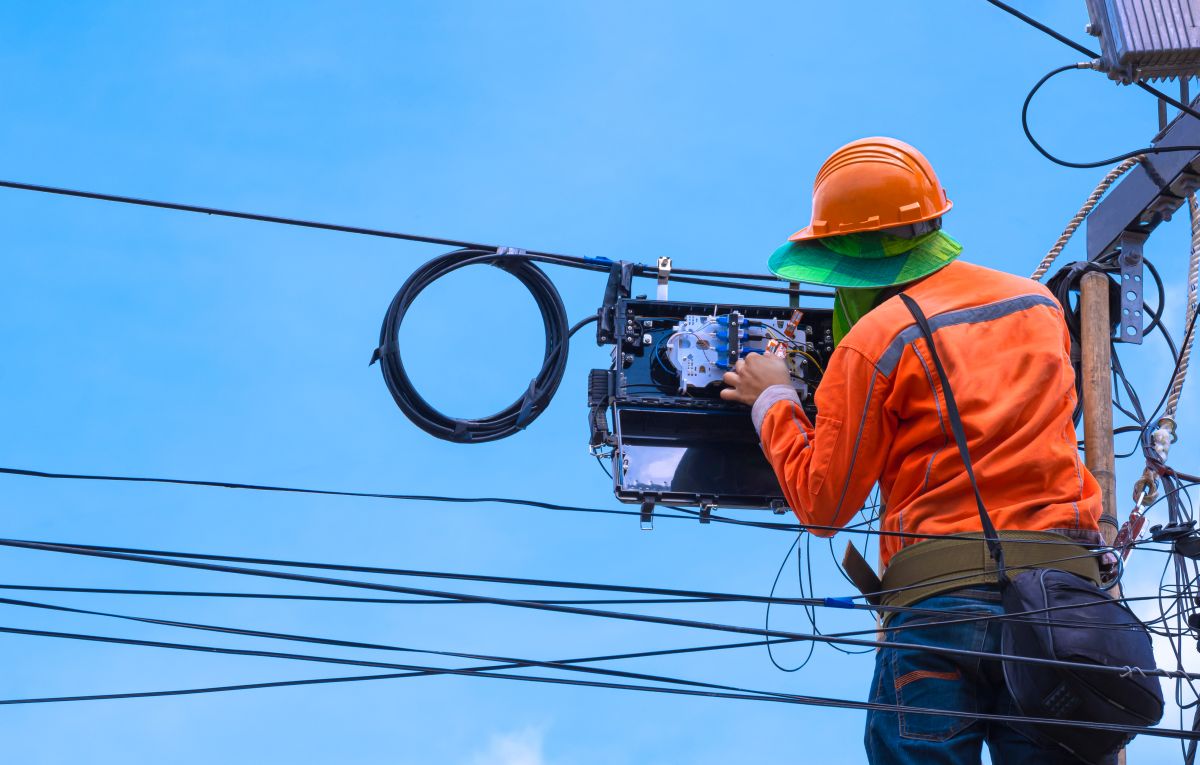Survey Forty-One Percent of Americans Are More Concerned with Power Outages - Certrec A utility worker, wearing an orange helmet and high-visibility jacket, is performing maintenance on high-voltage electrical equipment on a utility pole. He is using specialized tools and handling wires, with a clear blue sky in the background.