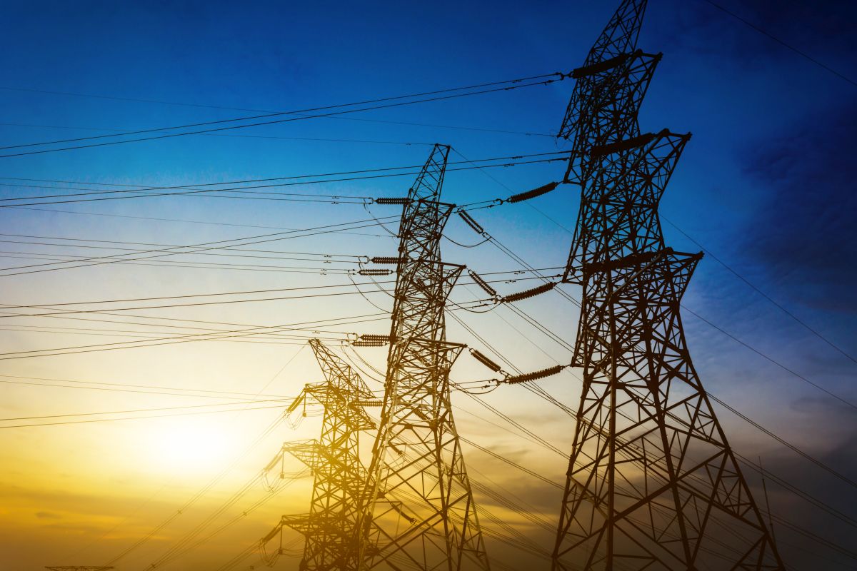 Silhouettes of electricity transmission pylons and power lines against a colorful sunset sky. The setting sun casts a golden glow behind the high voltage towers, highlighting energy infrastructure at dusk.