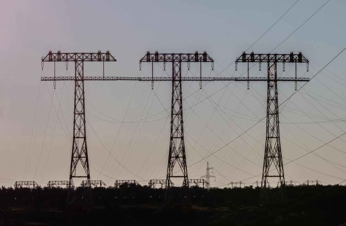 Three tall metal transmission towers carry multiple high voltage power lines stretching across the sky at dusk. The cables hang in wide curves between the structures, silhouetted above a dark tree line in the foreground.
