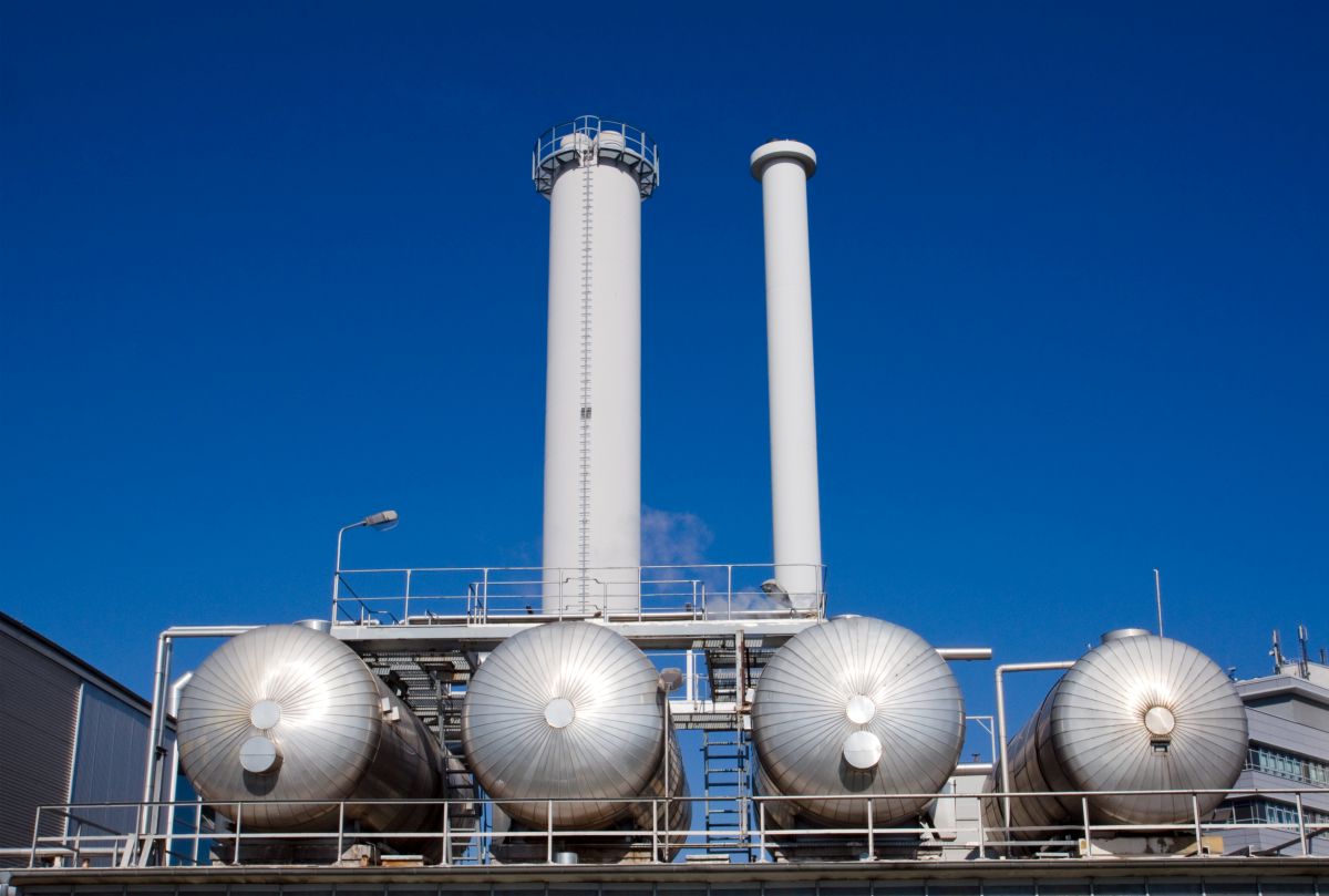 Industrial facility with four large cylindrical storage tanks aligned horizontally in the foreground and two tall white smokestacks rising behind them against a clear blue sky. Metal pipes, railings, and a ladder connect the structures, indicating a gas or energy processing plant.