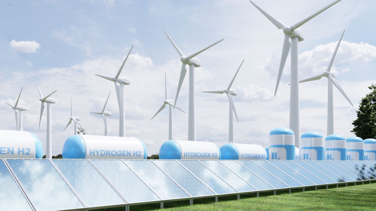 Hydrogen energy storage gas tank for clean electricity solar and wind turbine facility Solar panels line the foreground of a renewable energy site with multiple wind turbines spinning in the background and large hydrogen storage tanks labeled "HYDROGEN H2" positioned between them.