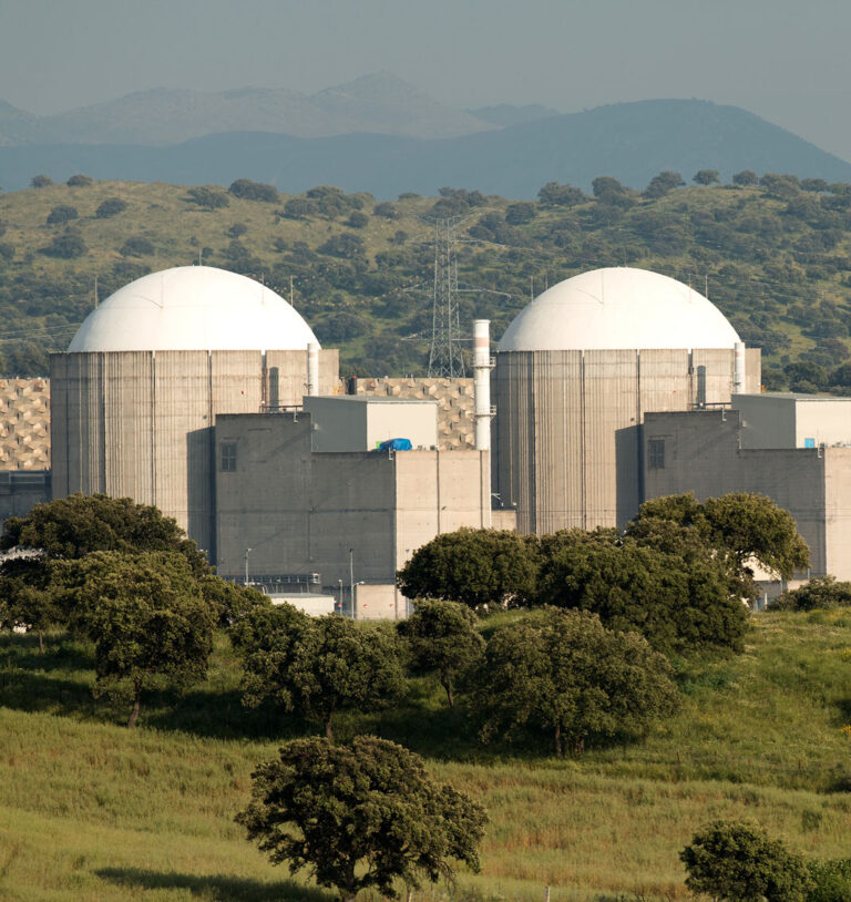 Two large nuclear reactor containment domes rising above concrete facility buildings with hills in the background. The image represents nuclear new builds and the infrastructure supporting next generation nuclear energy projects.