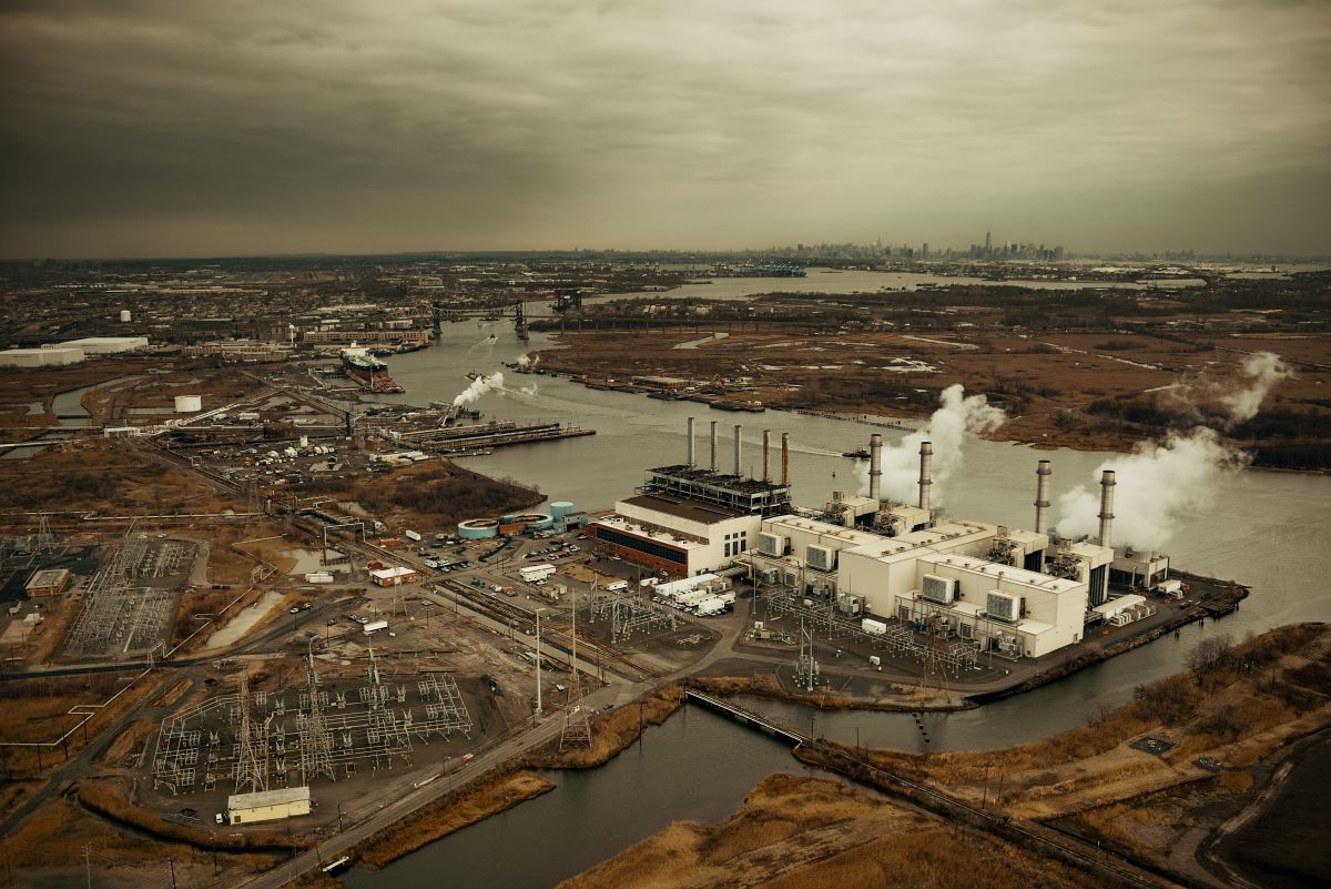 Manhattan aerial Aerial view of Manhattan with a large power plant along the waterfront, smokestacks releasing steam into an overcast sky. Industrial buildings, transmission equipment, and waterways fill the foreground, with the distant New York City skyline visible on the horizon.