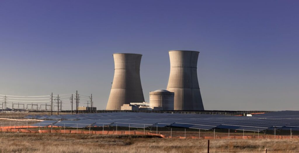 Nuclear power plant cooling towers rising behind rows of solar panels in California under a clear blue sky. The contrast between nuclear energy infrastructure and renewable solar arrays highlights the evolving mix of power generation sources.