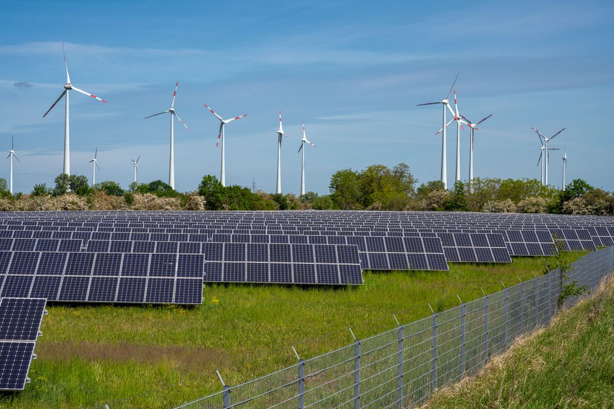 Solar panels in the foreground with wind turbines in the background, set against a clear sky and green landscape.