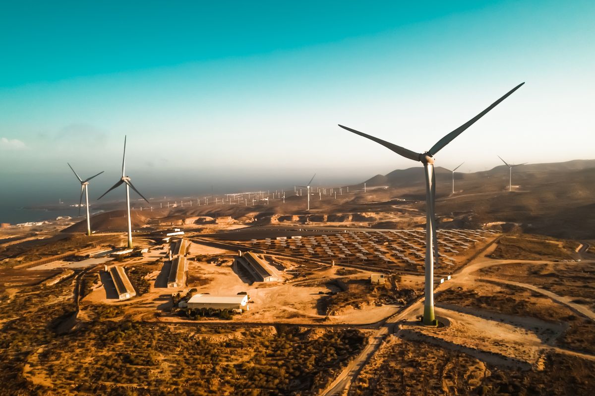 Panoramic view of wind turbines in the foreground with rows of solar panels in the background under a clear sky.