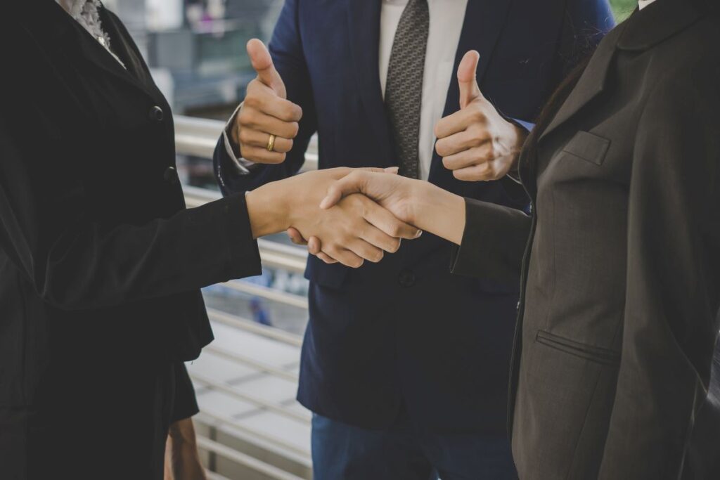 Two business people shaking hands to seal a deal, with another person giving a thumbs-up in the background.