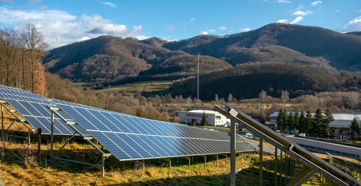 Solar panels on the meadow near the forest Green energy concept Rows of solar panels installed on a grassy hillside near a forest with mountains in the background and a small building nearby, showing renewable energy use in a rural landscape.