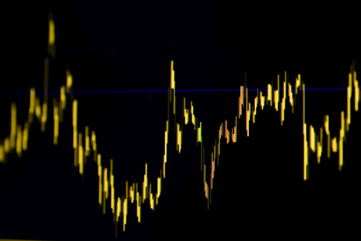 Bright yellow fluctuating stock chart line on a dark screen with a blue horizontal reference line, showing market price movements on a digital trading display.
