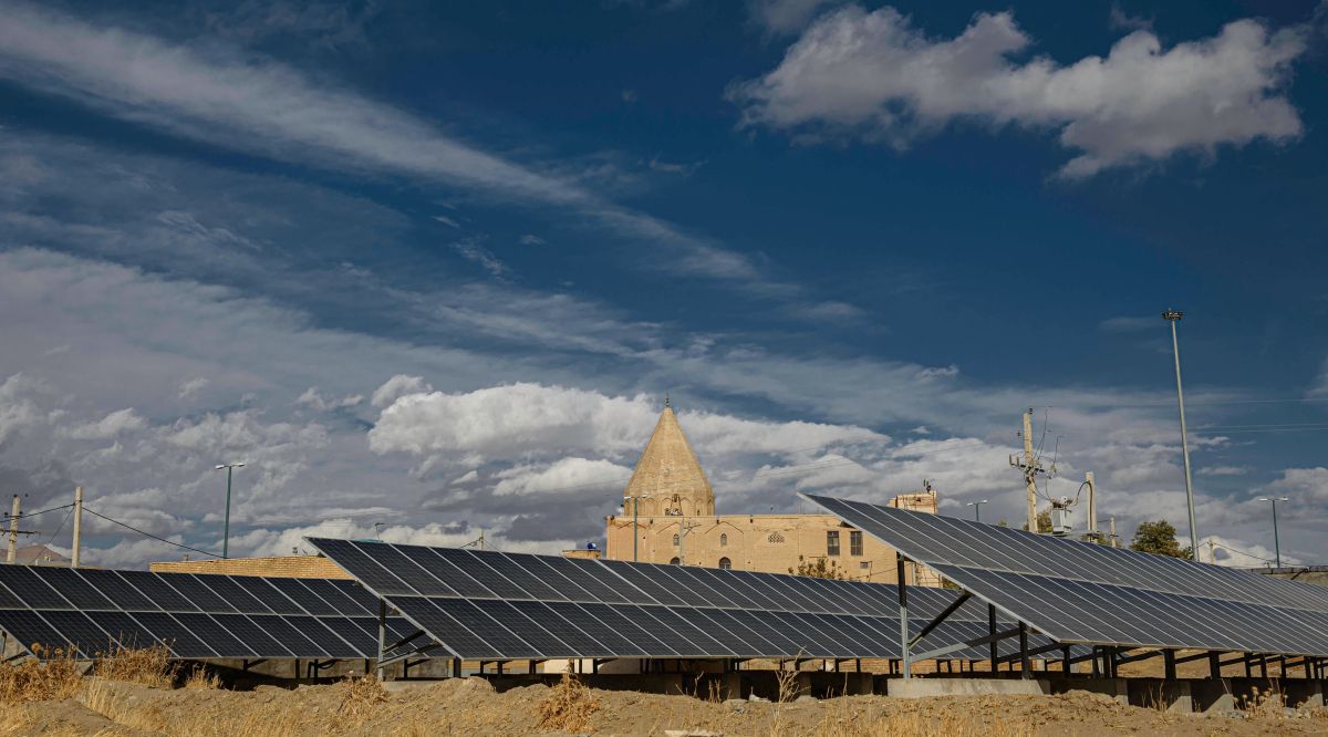 Solar panels installed in the foreground with a historic stone building and tower behind them under a partly cloudy sky, showing modern renewable energy alongside traditional architecture.