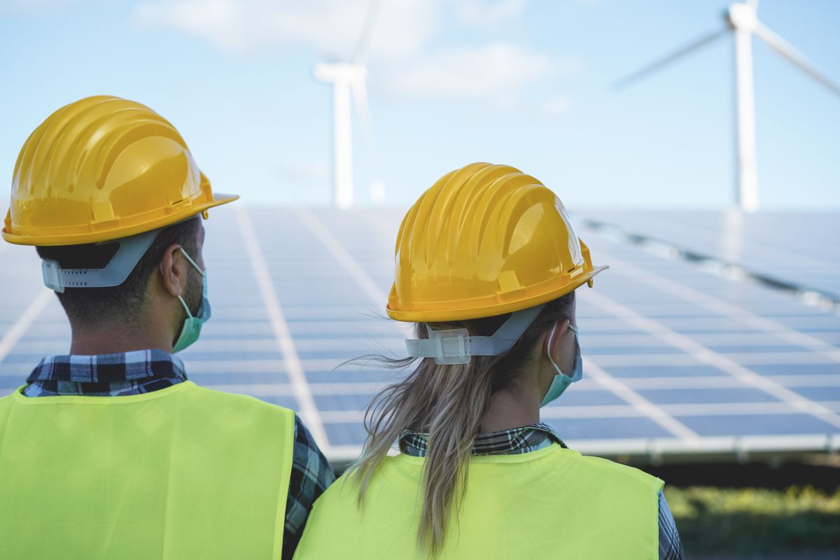 The Clean Energy Workforce Needs People Heres How a New Initiative Intends to Help - Certrec Two clean energy workers wearing yellow hard hats and safety vests stand in front of solar panels with wind turbines in the background. The image highlights the growing clean energy workforce supporting renewable power generation projects.