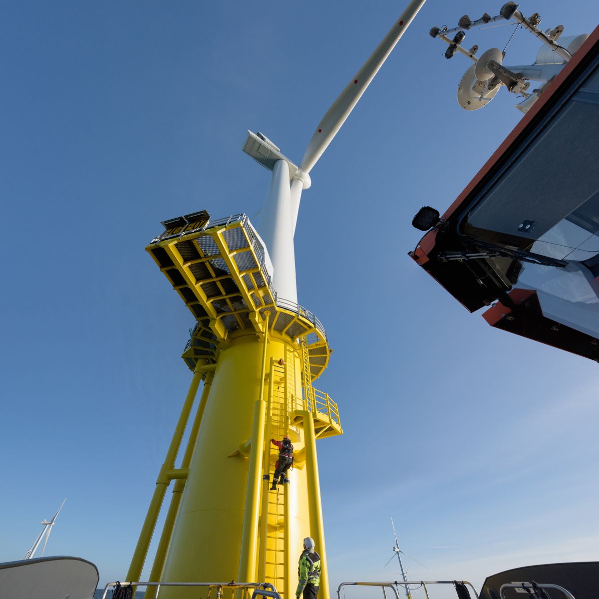 Low angle view of an offshore wind turbine tower beside a service boat under a clear blue sky.