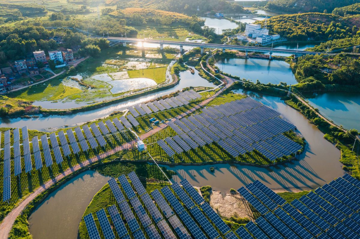 Aerial view of a solar power plant with rows of photovoltaic panels installed along winding waterways and green landscape. The image highlights large scale solar energy generation integrated into the surrounding environment.