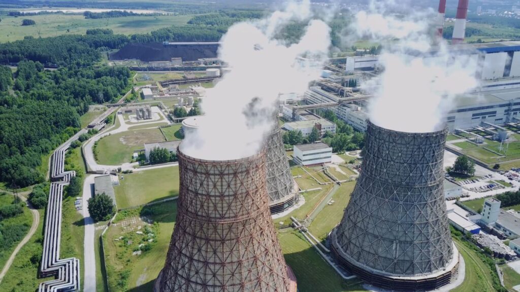 Wide aerial view of a nuclear power plant with cooling towers emitting steam, surrounded by green fields and infrastructure