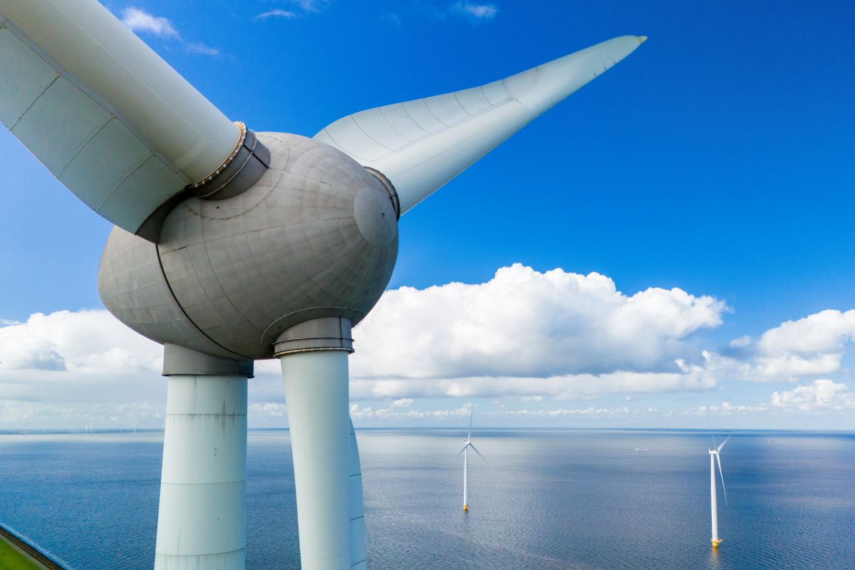Close view of a large offshore wind turbine with blades extending over the sea and additional turbines in the distance under a blue sky, highlighting renewable energy generation in coastal waters.
