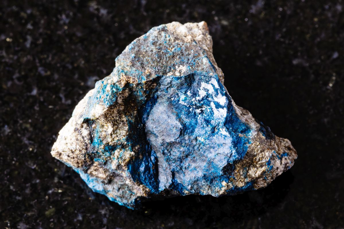 Rough piece of aerinite mineral with blue and white crystalline textures embedded in dark rock, displayed on a black granite surface to highlight its natural color and structure.