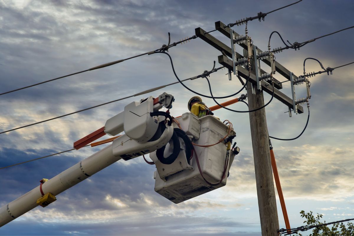 Utility worker in a bucket lift repairing power lines on a high voltage utility pole under a cloudy sky, showing maintenance work on electrical infrastructure after storm damage.