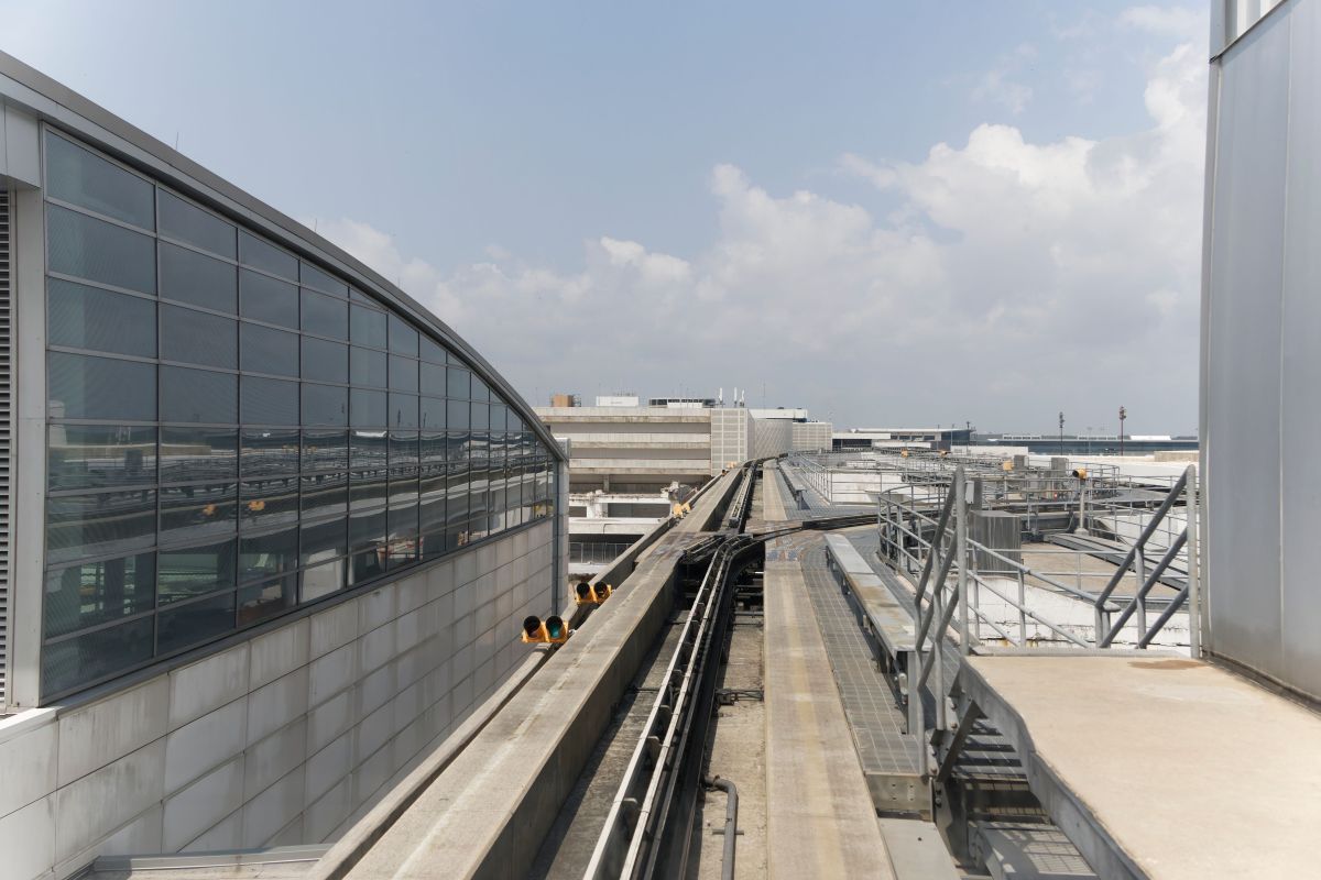 Elevated AirTrain tracks running alongside a modern glass terminal building with airport infrastructure in the background under a partly cloudy sky, showing urban transit at an airport.