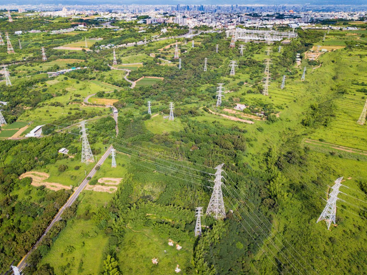 Aerial view of green landscape with multiple high voltage transmission towers and power lines stretching across fields toward a distant city, showing large scale electricity infrastructure connecting urban and rural areas.