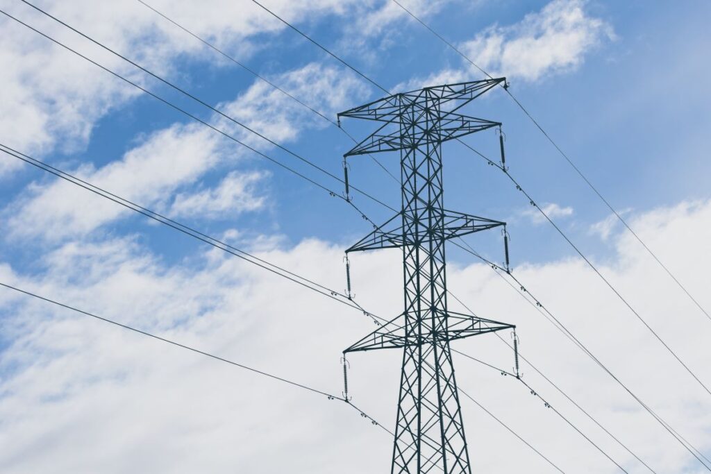 The image depicts an electric power transmission tower with a blue sky and scattered clouds in the background. The transmission lines are clearly visible, highlighting the infrastructure used to transport electricity.