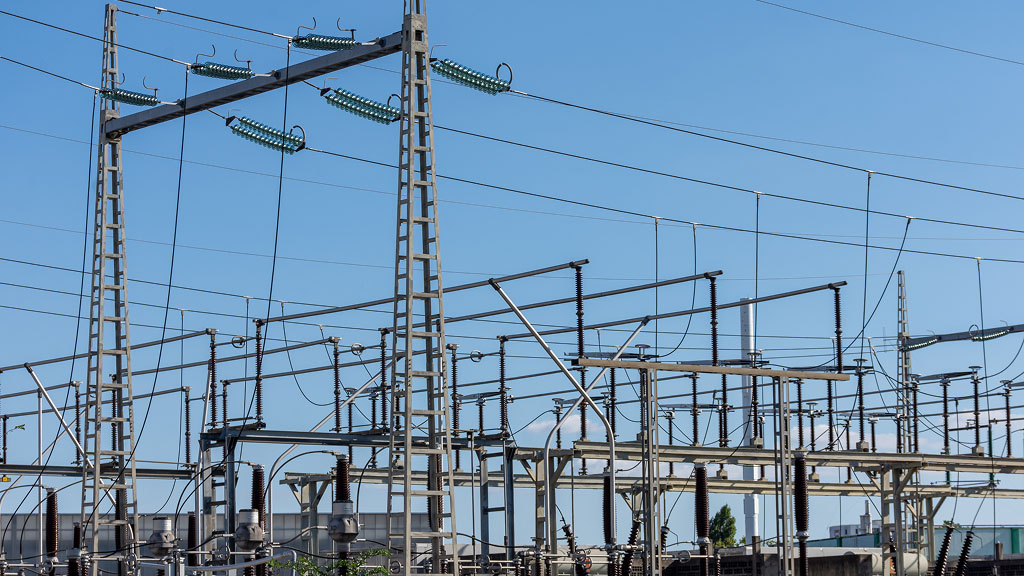 Electrical substation with overhead transmission lines, insulators, circuit breakers, and protection equipment against a clear blue sky. The high voltage infrastructure highlights reliable protection system maintenance and compliance with NERC PRC-005 standards for grid reliability.