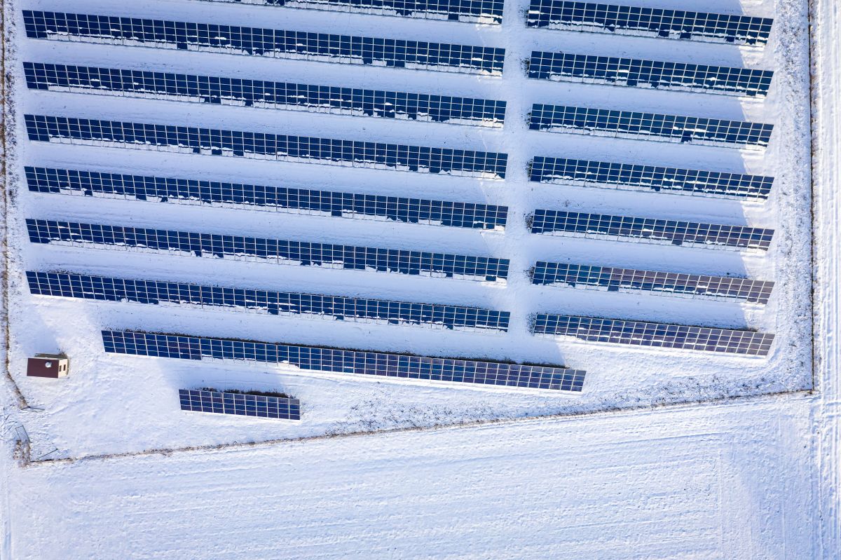 Aerial view of rows of solar panels partially covered in snow across a white field, showing a photovoltaic farm operating in winter conditions.